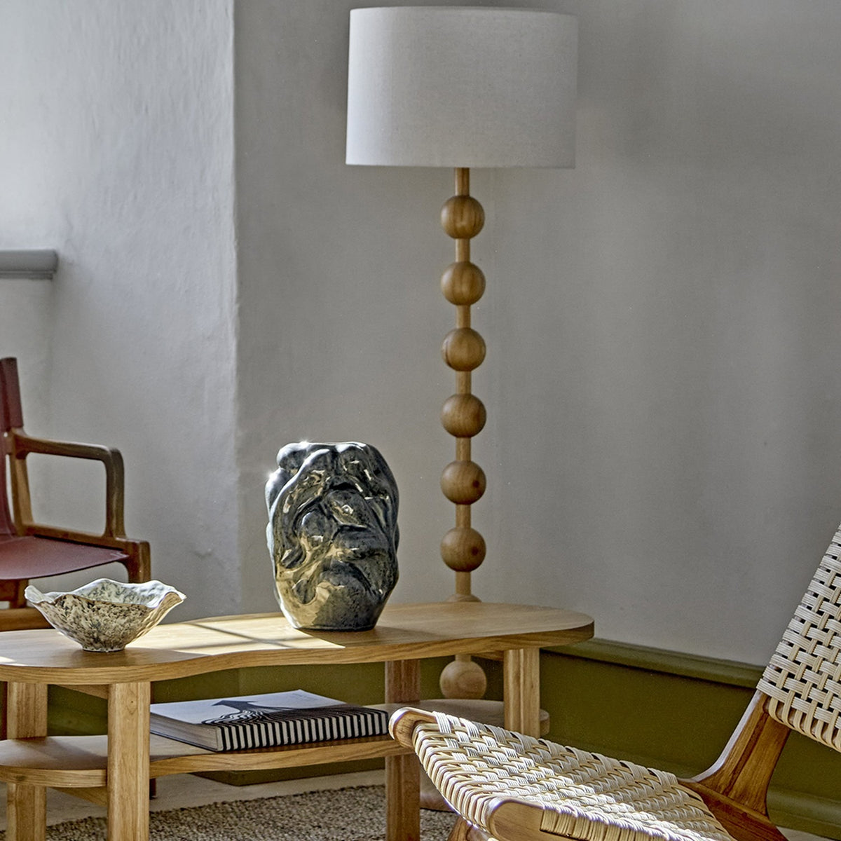 Giacoma Wooden Floor Lamp in natural wood with a stacked sphere stem, round base and linen drum shade, styled in a living room beside a coffee table and woven chair.