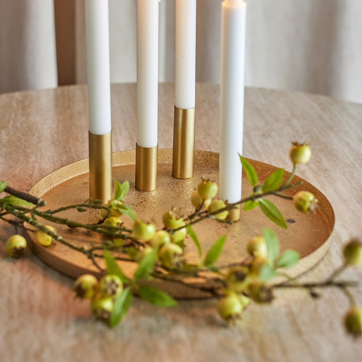 Maxime Iron Christmas Candle Holders Set in gold, round textured tray with slim tube holders and white taper candles, styled with green berry foliage on a wooden table.