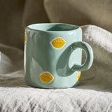 Agnes Stoneware Cup in green, a glossy ceramic mug with a curved handle and hand-painted yellow lemon motifs, shown on a neutral linen fabric background.