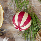 Wynn Striped Christmas Bauble in red and cream, a fabric-wrapped ball with a beaded gold hanging loop, shown hanging from a pine branch on a wooden surface.