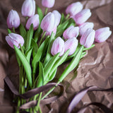 Artificial bouquet of pink tulips with closed, cupped blooms and long green stems, tied with ribbon on a brown paper surface.