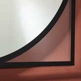Corner detail of the Circo square wall mirror, showing the black frameless edge and partially transparent printed glass over a terracotta wall.