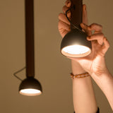 Berry S Aluminium LED pendant light in matte black, hand adjusting the domed, angled head on a slim tube suspension, with a second illuminated pendant visible in the background.