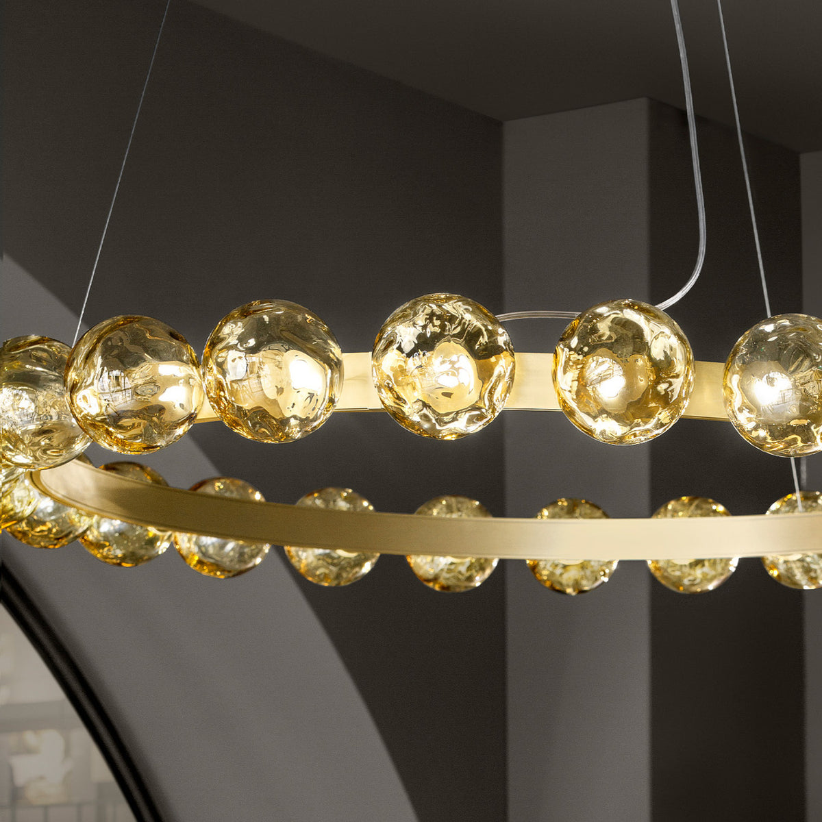 Murané R 25 ring chandelier, 110cm brushed champagne band set with caramel glass spheres, textured amber globes catching light against a grey ceiling backdrop.