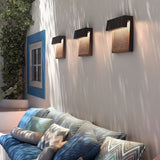 Three Sisal A/01 LED outdoor wall lights with brown woven fibre front panels and angled hoods, mounted above a patio sofa with cushions and a blue-framed window.