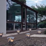 Cornet B/27 Cone LED bollard lights in Stone Grey, tapered cone bases with wavy stacked layers, lining a garden path beside a glass-fronted building at dusk.