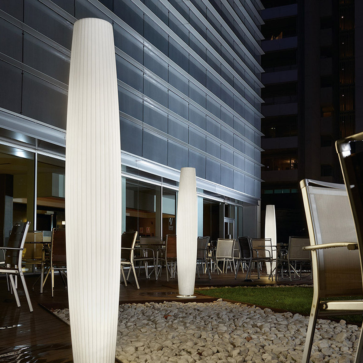 Maxi P/180 Cylinder LED Outdoor Floor Lamp in white polyethylene with ribbed texture, illuminated on a terrace among outdoor chairs and gravel landscaping at night.