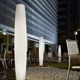 Maxi P/180 Cylinder LED Outdoor Floor Lamp in white polyethylene with ribbed texture, illuminated on a terrace among outdoor chairs and gravel landscaping at night.