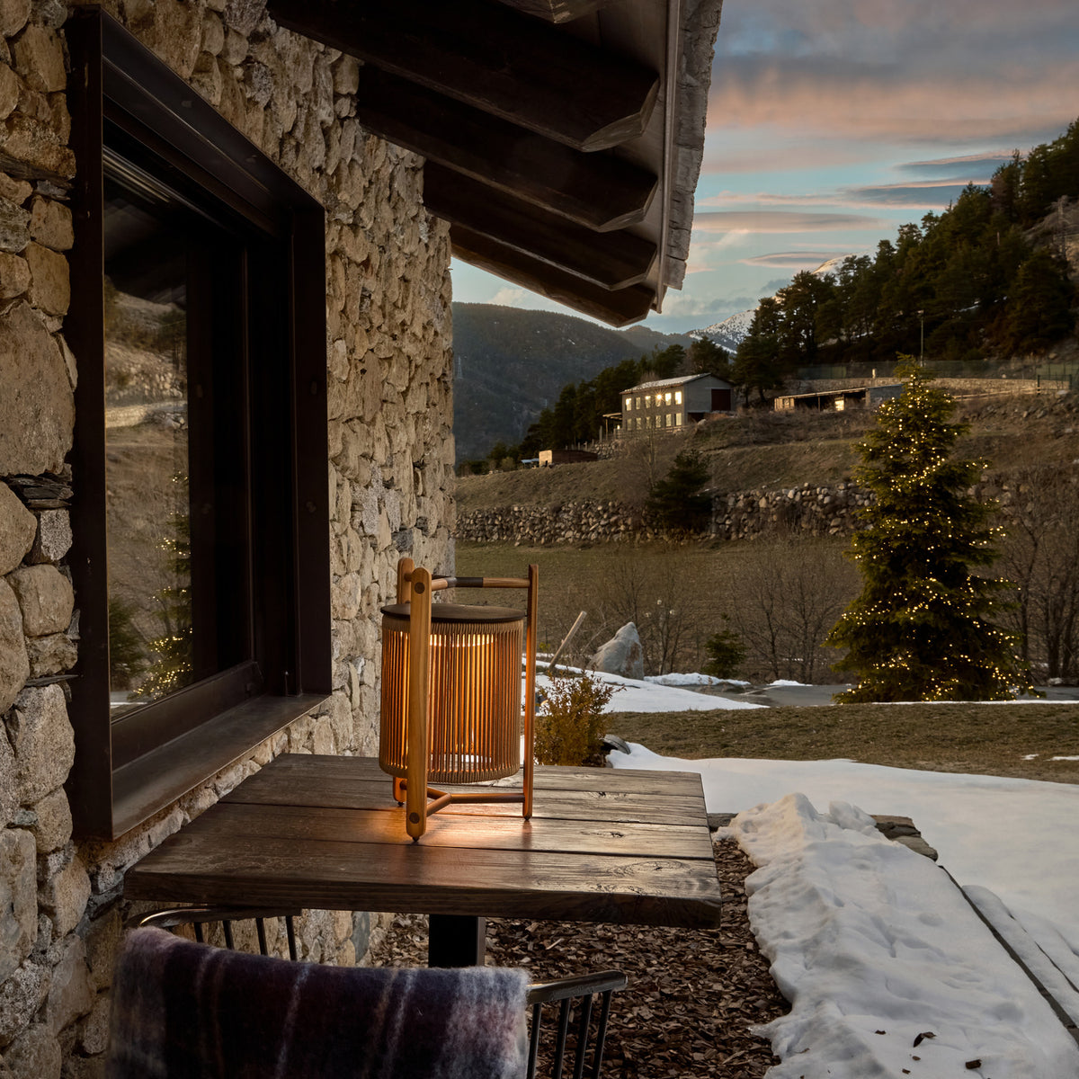 Kando M/40/R rechargeable outdoor table lamp with sand nautical cord cylinder and teak wood frame, glowing on a wooden table against a snowy mountain terrace at dusk.