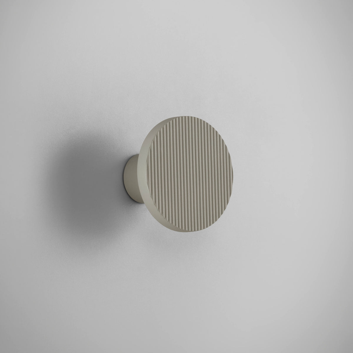 Arid A/01 round aluminium LED wall light in Textured Pebble Grey, fluted disc face on a short cylindrical stem, mounted on a plain light grey wall.