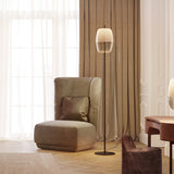 Adamas Smoke Grey Glass LED Floor Lamp with faceted honeycomb-patterned shade on a slim black stem and round base, shown beside an armchair in a living room setting.