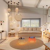 Nans Bag/38 outdoor floor lamp with brown woven lattice drum shade and teak carry handles, shown glowing orange on a round rug in a living room with hanging pendant lights.