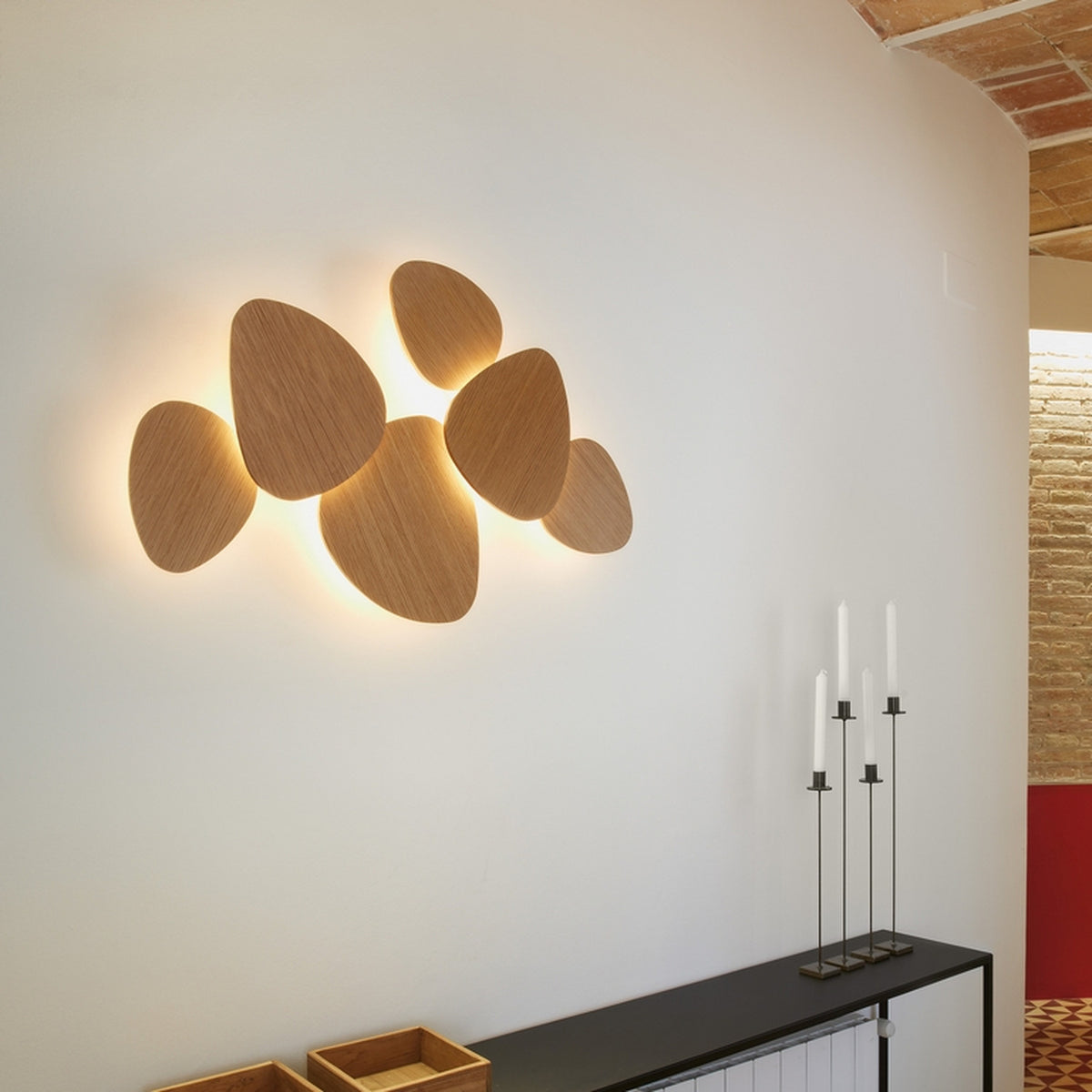 Tria 6 Light Wood LED Wall Light in natural oak, six pebble-shaped panels clustered together and backlit against a white wall, shown above a console table with candlesticks.