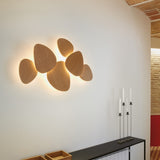 Tria 6 Light Wood LED Wall Light in natural oak, six pebble-shaped panels clustered together and backlit against a white wall, shown above a console table with candlesticks.