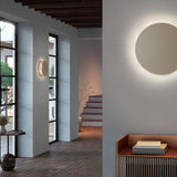 Helios A/02 round LED wall light in Oak Brown & Beige fabric finish, shown glowing softly on a hallway wall and as a backlit disc beside an entryway console.