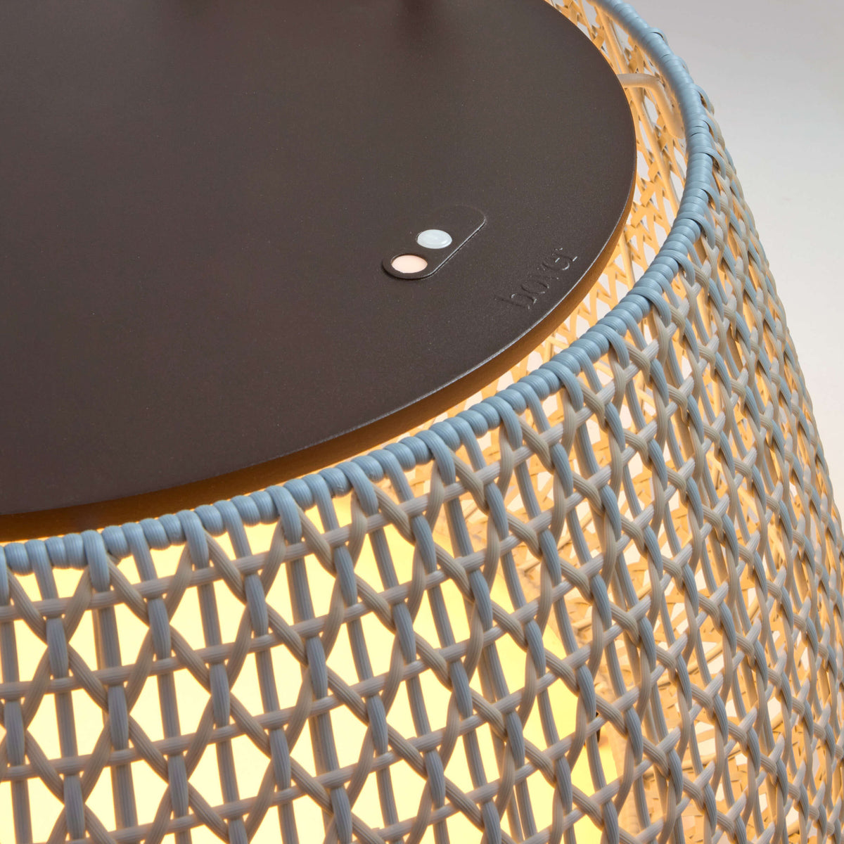 Close-up of the Nans Bag/38 outdoor floor lamp showing its flat dark top with control buttons and the beige open woven shade glowing from the integrated LED inside.