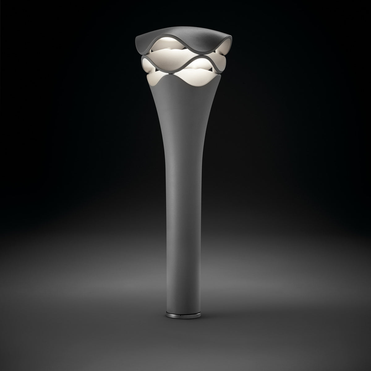 Cornet B/77 tiered LED bollard light in stone grey aluminium, with a slender flared stem rising to a wavy layered head glowing warm white, shown on a dark background.
