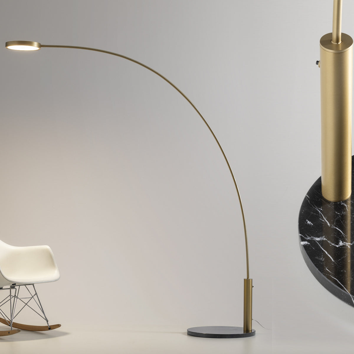 Dip Nero Marquina arc LED floor lamp with a slim brushed brass curved arm, round disc head and black marble base, shown beside a white rocking chair on a plain background.