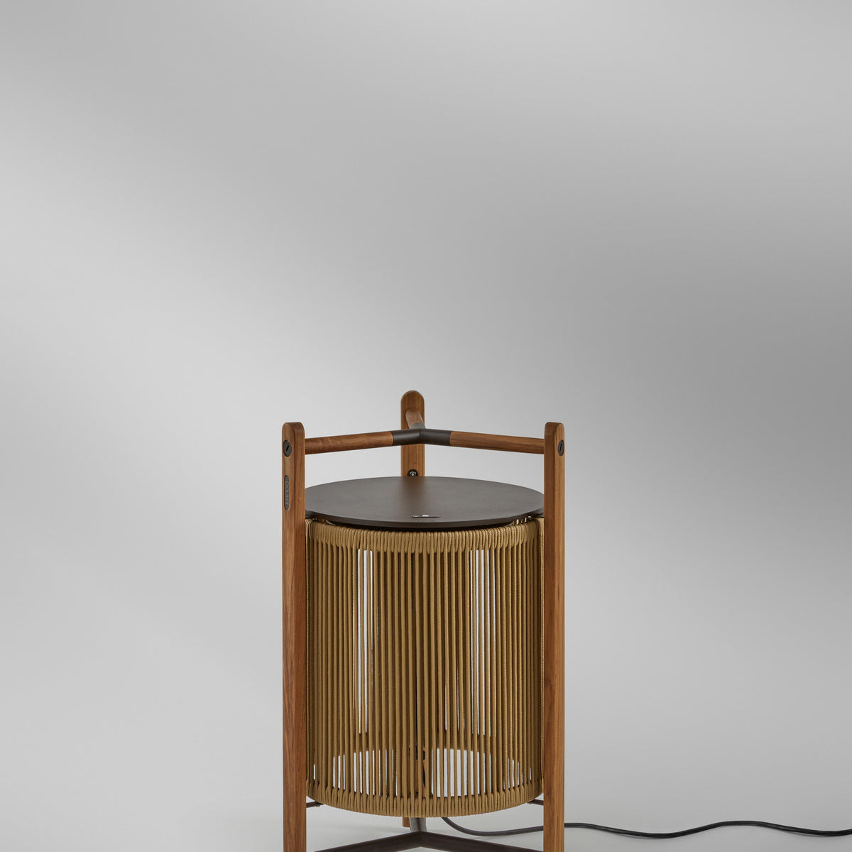 Kando P/76 outdoor floor lamp with sand-coloured nautical cord cylinder in a three-legged teak wood frame, dark round lid and wooden handle, shown against a plain light grey background.