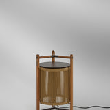 Kando P/76 outdoor floor lamp with sand-coloured nautical cord cylinder in a three-legged teak wood frame, dark round lid and wooden handle, shown against a plain light grey background.