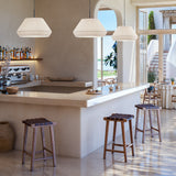 Mei 38 Ribbon Cone Pendant Lights in white pleated ribbon shades, hung in three sizes above a bar counter with wooden stools in a sunlit dining space.