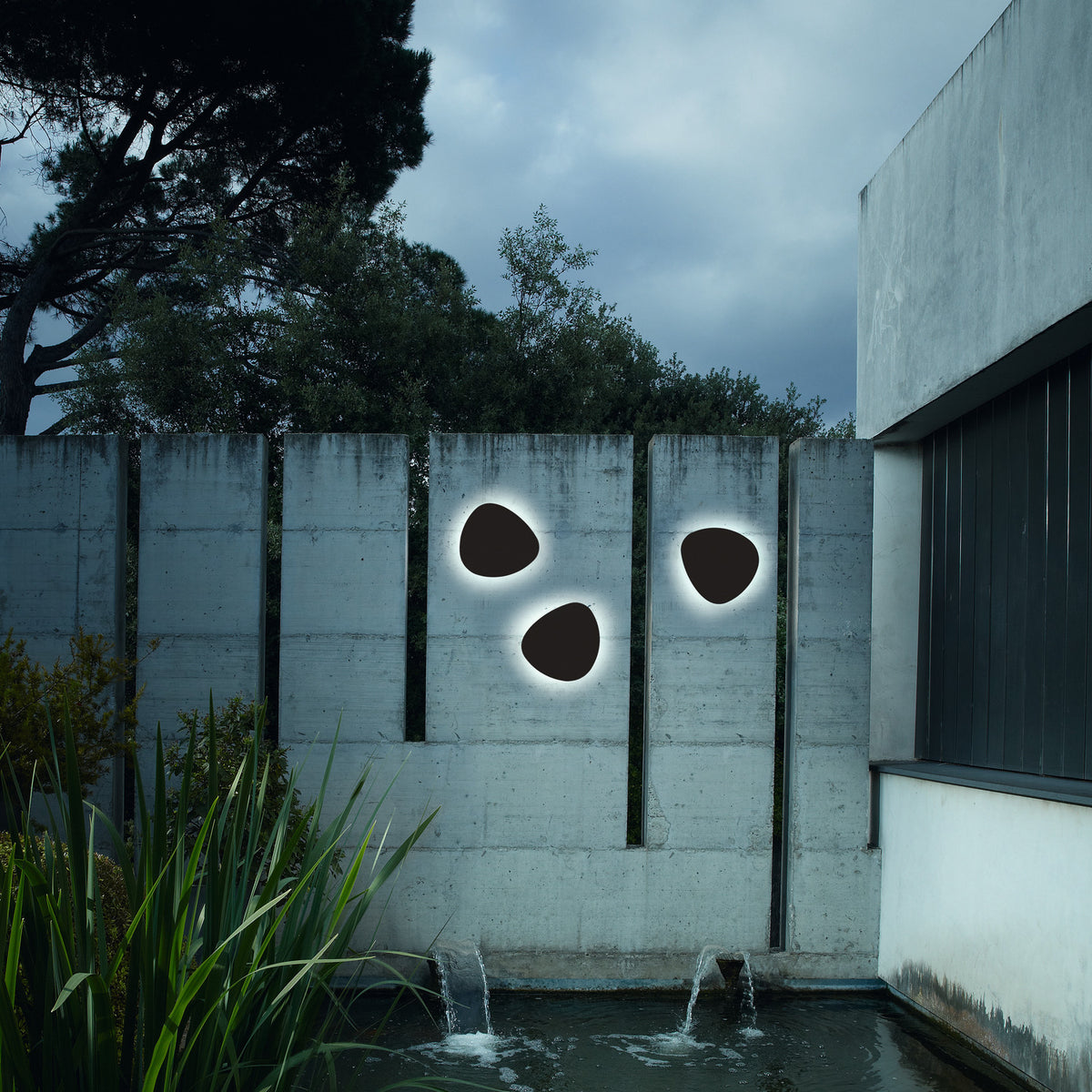 Three pebble-shaped Tria 04 LED outdoor wall lights in graphite brown metal mounted on a concrete garden wall, each glowing with a soft warm-white halo.
