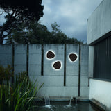 Three pebble-shaped Tria 04 LED outdoor wall lights in graphite brown metal mounted on a concrete garden wall, each glowing with a soft warm-white halo.