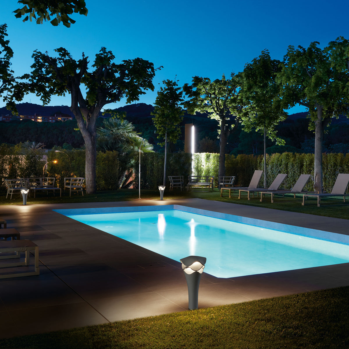 Cornet B/52 Cone LED Bollard Light in stone grey, illuminated poolside at night, with its wavy white cap glowing softly amid a garden and lounge seating.