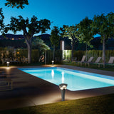 Cornet B/52 Cone LED Bollard Light in stone grey, illuminated poolside at night, with its wavy white cap glowing softly amid a garden and lounge seating.