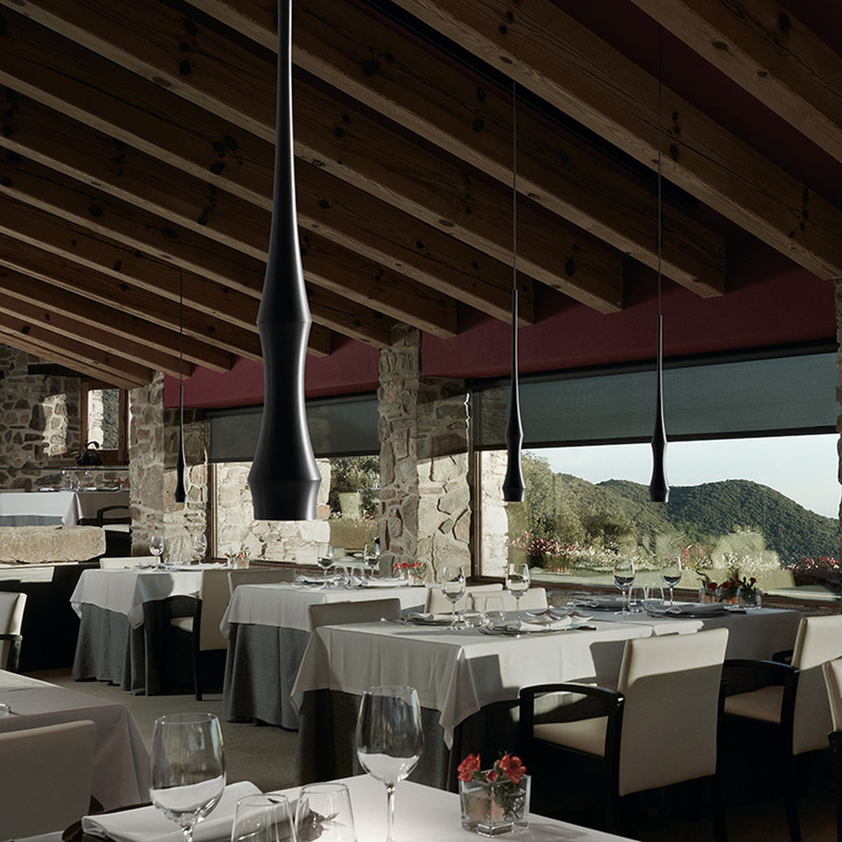 Slend 01 aluminium pendant lights in matte black with slim, tapering waisted bodies, hung above restaurant dining tables with white linens beneath exposed wood beam ceilings.