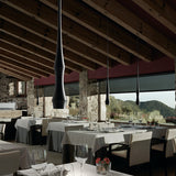 Slend 01 aluminium pendant lights in matte black with slim, tapering waisted bodies, hung above restaurant dining tables with white linens beneath exposed wood beam ceilings.