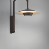Fanalet A/02 outdoor wall light with a tall arched arm in graphite brown and a wide flat shade with white underside, illuminated against a plain grey background.