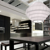 Mos 03 Ribbon Tiered Pendant Light with a large rounded shade of pleated white translucent ribbon rings, shown lit above a dark dining table in a modern kitchen setting.