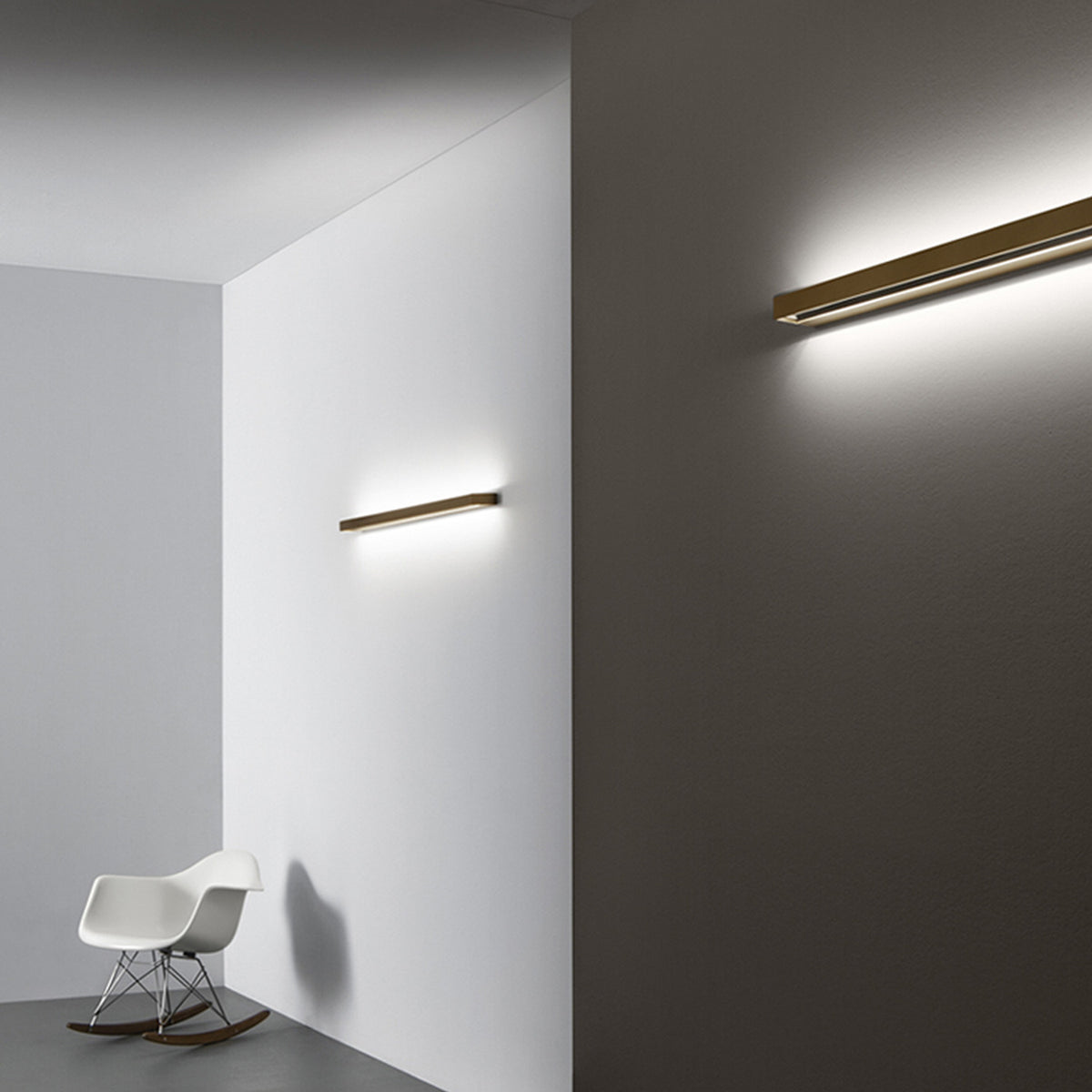 Zero Line Metal Linear LED Wall Light, 52cm brass bar fixture shown on a grey and white wall in a room with a white rocking chair, casting warm linear light.
