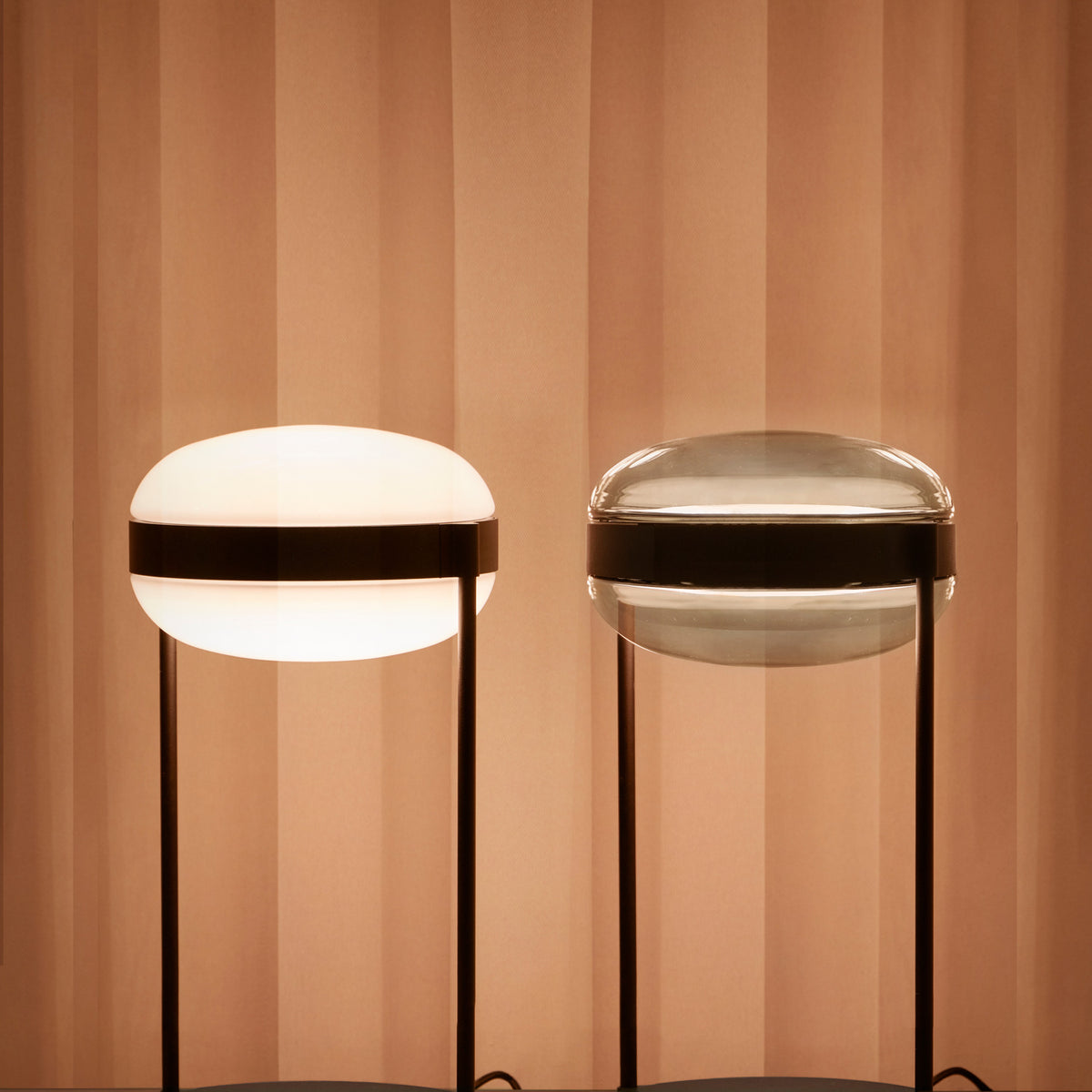 Beaded T Glass LED Table Lamp shown in Opal White (lit) and Smoke Grey versions, rounded flattened sphere shades with dark centre bands on slim twin-rod stands, set against a warm pleated backdrop.
