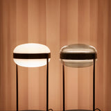Beaded T Glass LED Table Lamp shown in Opal White (lit) and Smoke Grey versions, rounded flattened sphere shades with dark centre bands on slim twin-rod stands, set against a warm pleated backdrop.