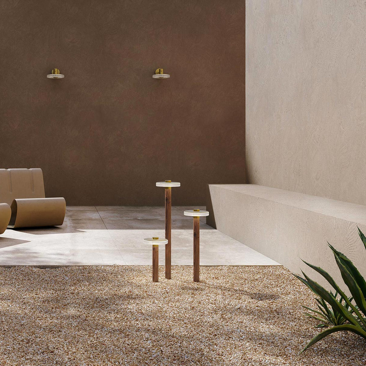 Venexia Glass LED Outdoor Floor Lamp in three heights, slim wood columns with round matt brass bases and glass discs, set on gravel in a courtyard.