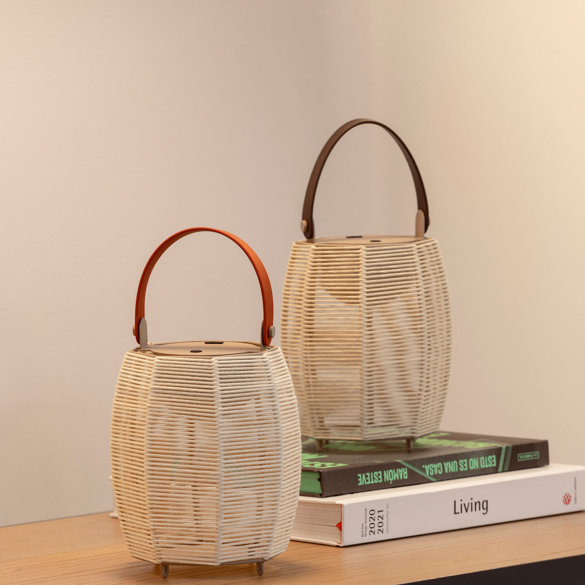 Tanit M/29/R rechargeable table lamp with woven beige lantern-style shade and brown curved carry handle, shown beside a larger matching lantern lamp on a wooden shelf with books.