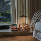 Valentina M/29 Table Lamp with wavy black and copper shade on a slim black stem, glowing on a wooden stool beside a stack of books in a bedroom setting.