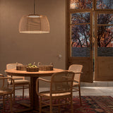 Tanit S/61 Fabric LED Pendant Light with wide faceted beige cotton shade on matt black stem, shown lit above a wooden dining table in a living room setting.