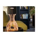 Domita M/36 wood bell table lamp with natural beech slatted shade and brown stem, lit on a side table in a living room with bookshelf.