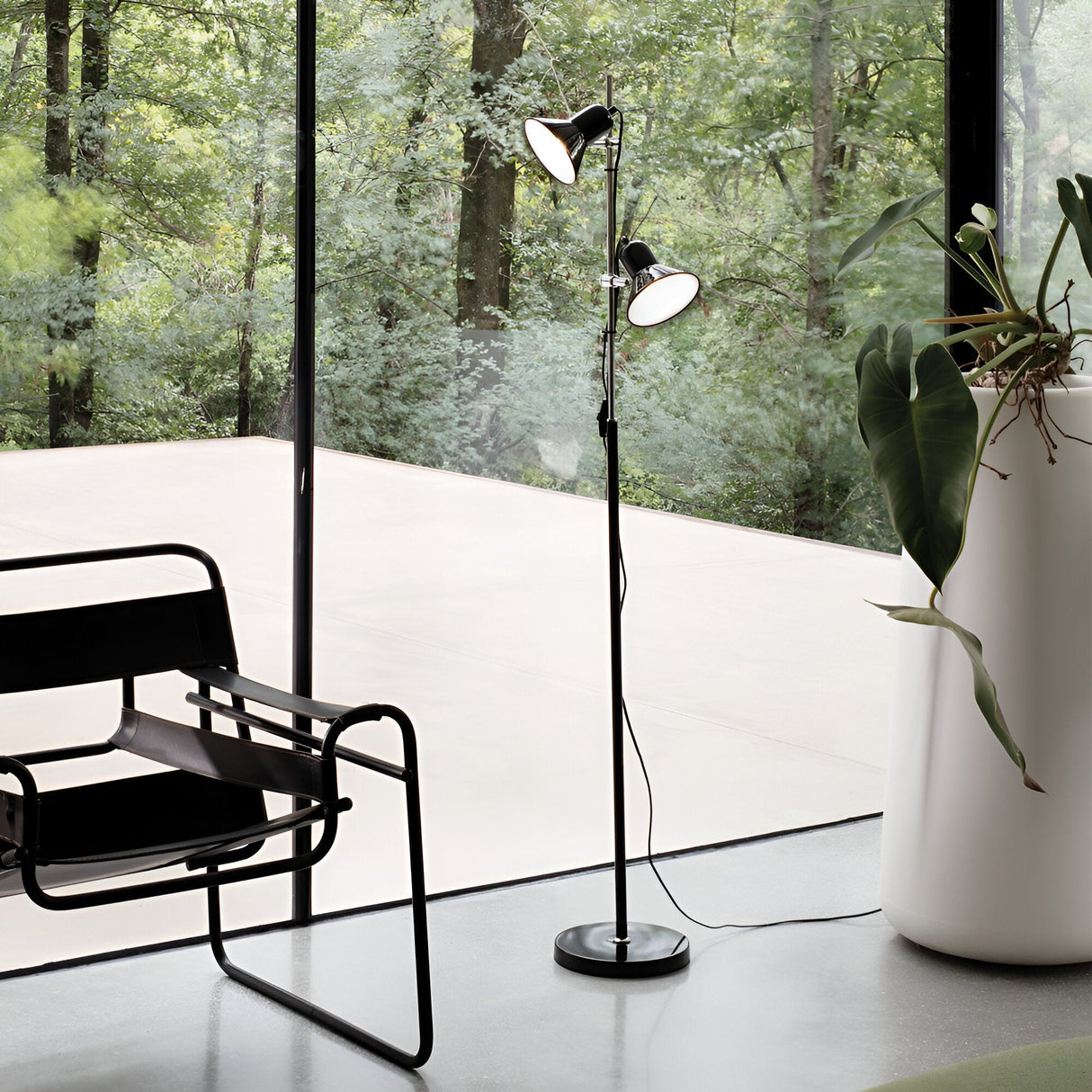 A modern living space highlights the Seraphic Floor Lamp - Black, known for its sleek design and adjustable diffusers, complemented by a black chair with a metal frame. A tall plant is positioned by the glass wall, providing calming views of the lush green forest in this bright and minimalistic setting.