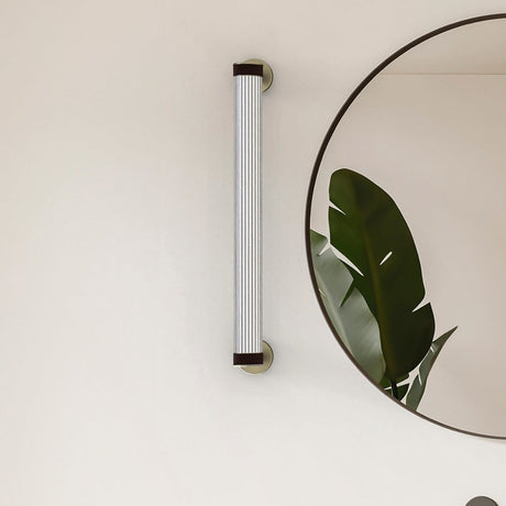 Roseline LED Ribbed Glass Linear Wall Light in black and beige, mounted vertically beside a round mirror with a trailing plant leaf, fluted glass bar held by beige straps on black circular bases.