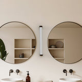 Roseline LED Ribbed Glass Linear Wall Light in gold and brown, mounted vertically between two round mirrors in a bathroom with open shelving and twin basins.