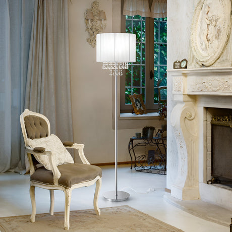 A cozy room with an ornate fireplace, armchair, and the Brillis Crystal Floor Lamp - Chrome. A window with sheer curtains reveals greenery outside, adding a touch of nature to the elegant, classical decor marked by luxury lighting.