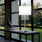A contemporary indoor scene with a large window overlooking a lush green lawn exudes a serene atmosphere. A sleek table showcases a bird sculpture alongside the Brillis Crystal Table Lamp - White, its shade embellished with cascading crystals. Sunlight pours in, creating gentle shadows.