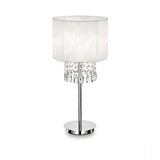 The Brillis Crystal Table Lamp - White boasts a sleek silver stand and round base complemented by a cylindrical white fabric shade. Below the shade, decorative crystal droplets enhance its modern design, adding an elegant touch to the ambient atmosphere.