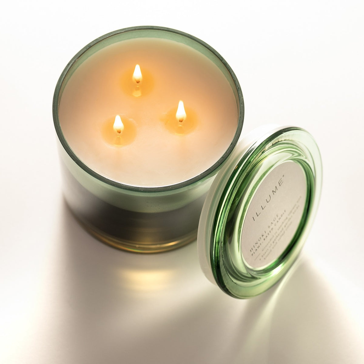 Hinoki Sage Glass Scented Candle 590g in a green glass jar with three lit cotton wicks, shown with its glass lid resting alongside against a plain white background.