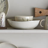 Bea Crackle Stoneware Deep Plate in cream with botanical leaf motif, nested inside a matching oval bowl on a shelf with a mug and wooden accent.