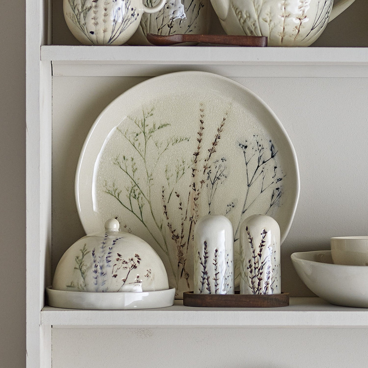 Bea Crackle Stoneware Dinner Plate in cream with floral grass motifs, displayed on a white shelf beside a matching cloche, shakers, and bowl.